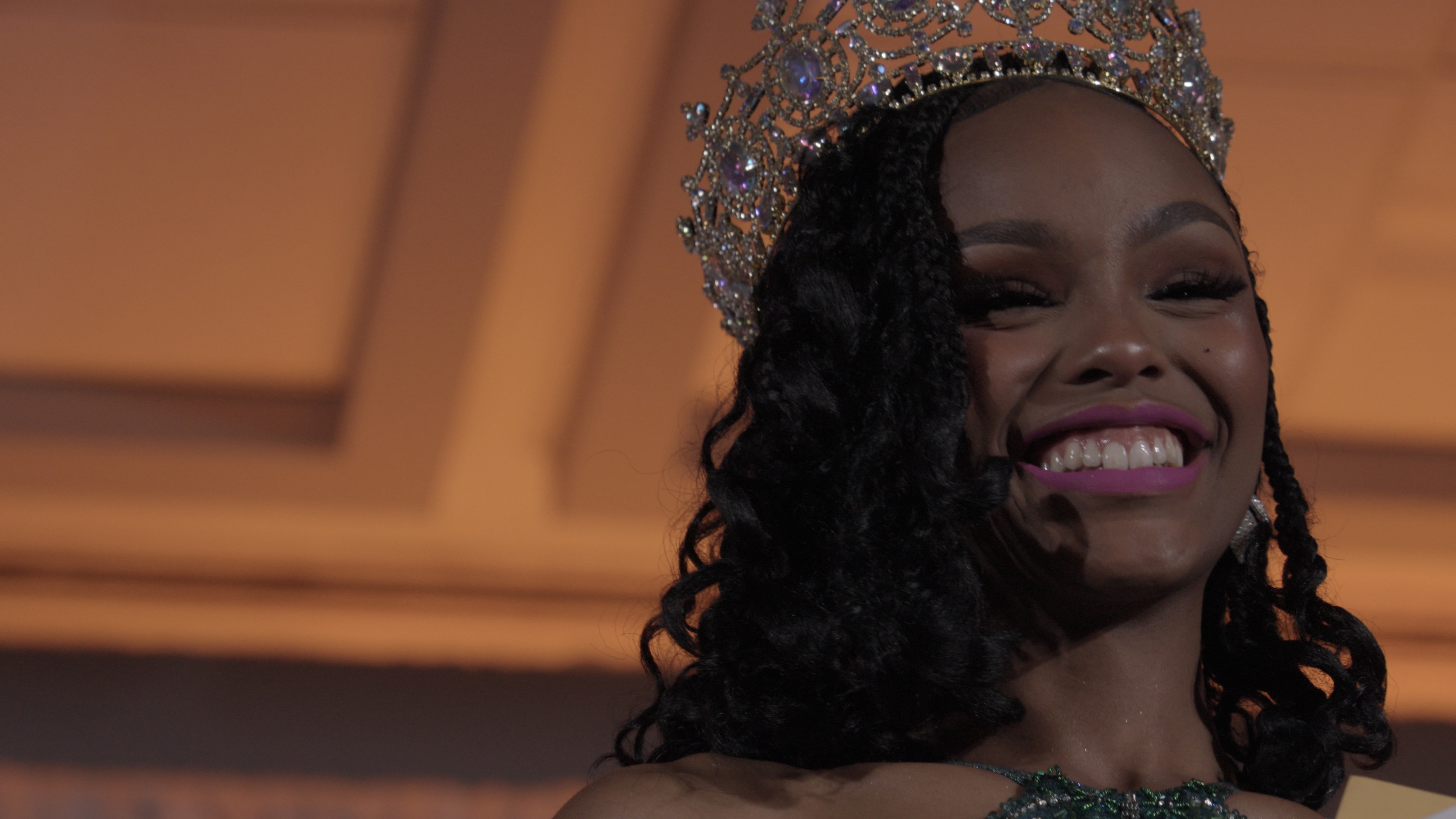 Woman in crown and green gown beaming with joy at pageant