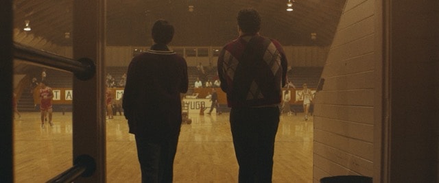 Two young men standing in a gymnasium tunnel watching a basketball game