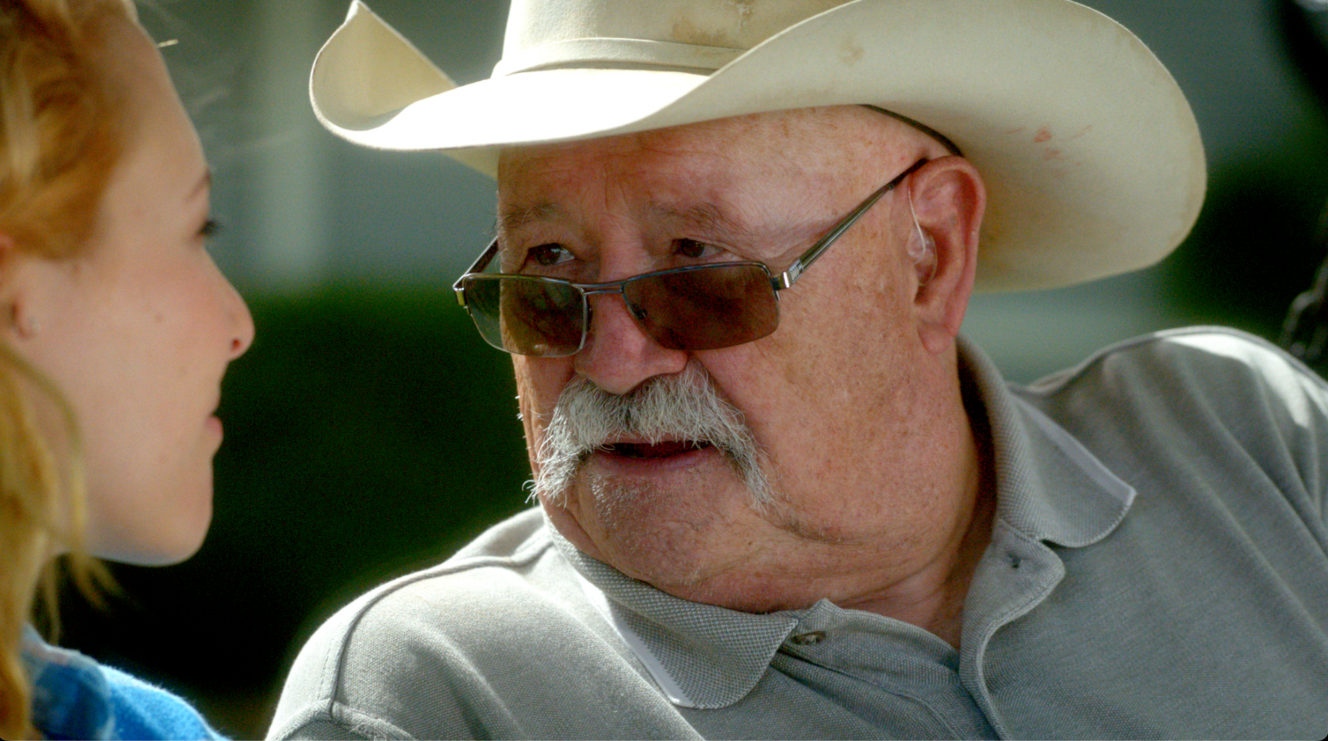 Barry Corbin in cowboy hat in close conversation with young woman