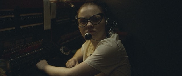 Woman in cat-eye glasses and headset at a radio control board