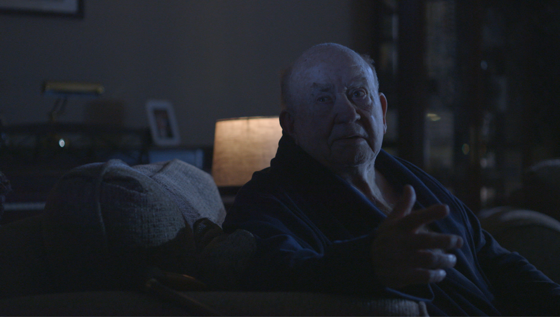 Elderly man in dim light speaking with expressive hands